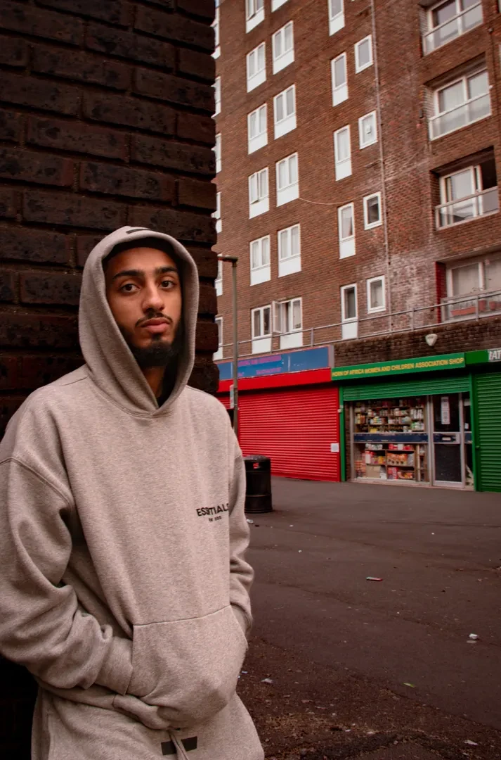 A young person in a hoodie is standing in an urban environment, looking at the camera with a neutral expression.