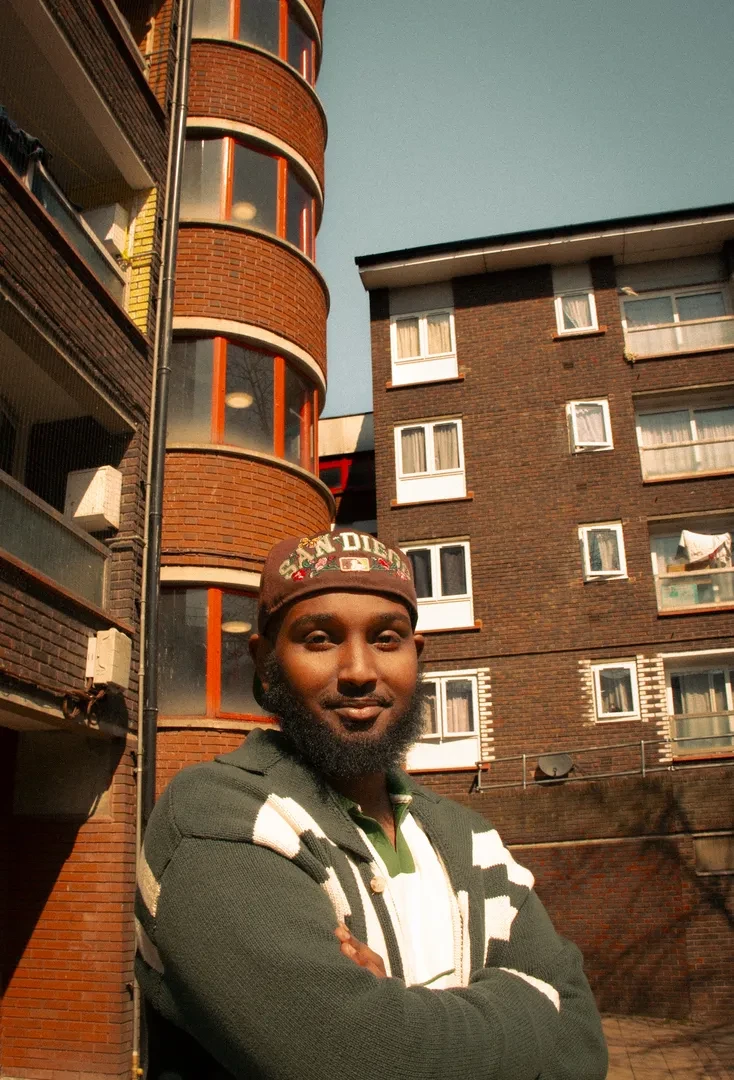 A young person bearded of color in an urban environment, looking at the camera with a smile.