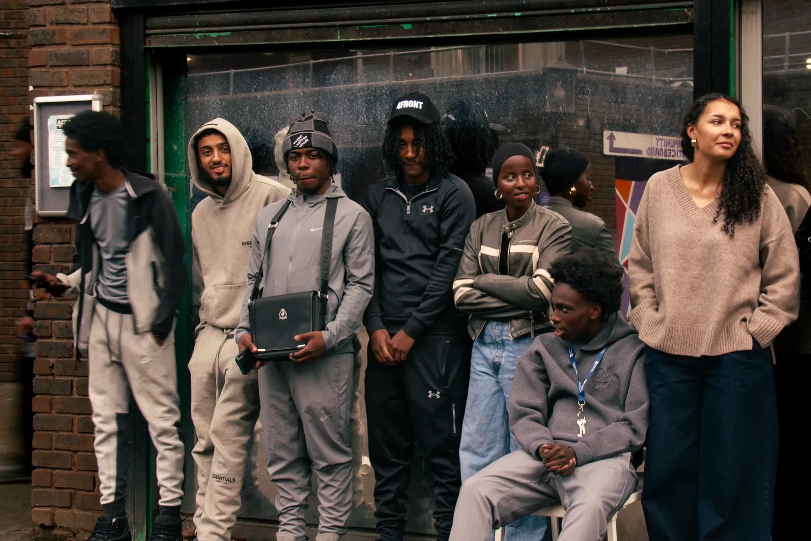 A group of seven young people of color stand and sit casually in front of a brick building.