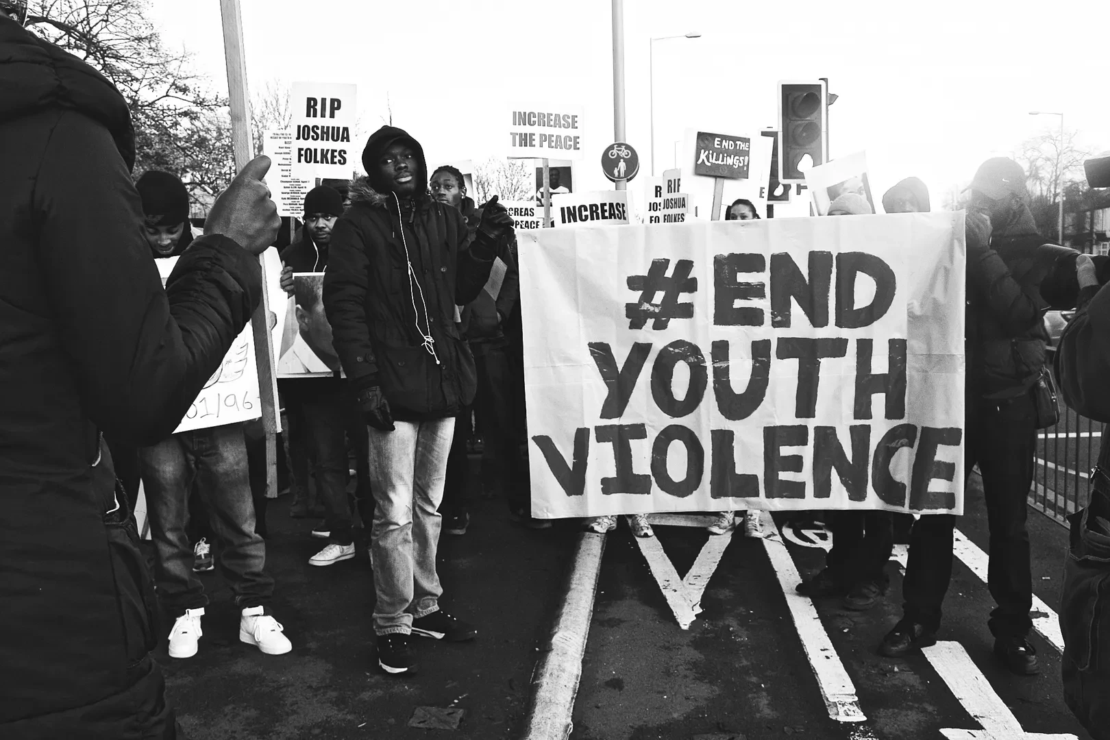 A black and white shot from a protest, where a young black person on the front stand next to a large sign that reads "# END YOUTH VIOLENCE". Many other people holding signs are visible in the background.