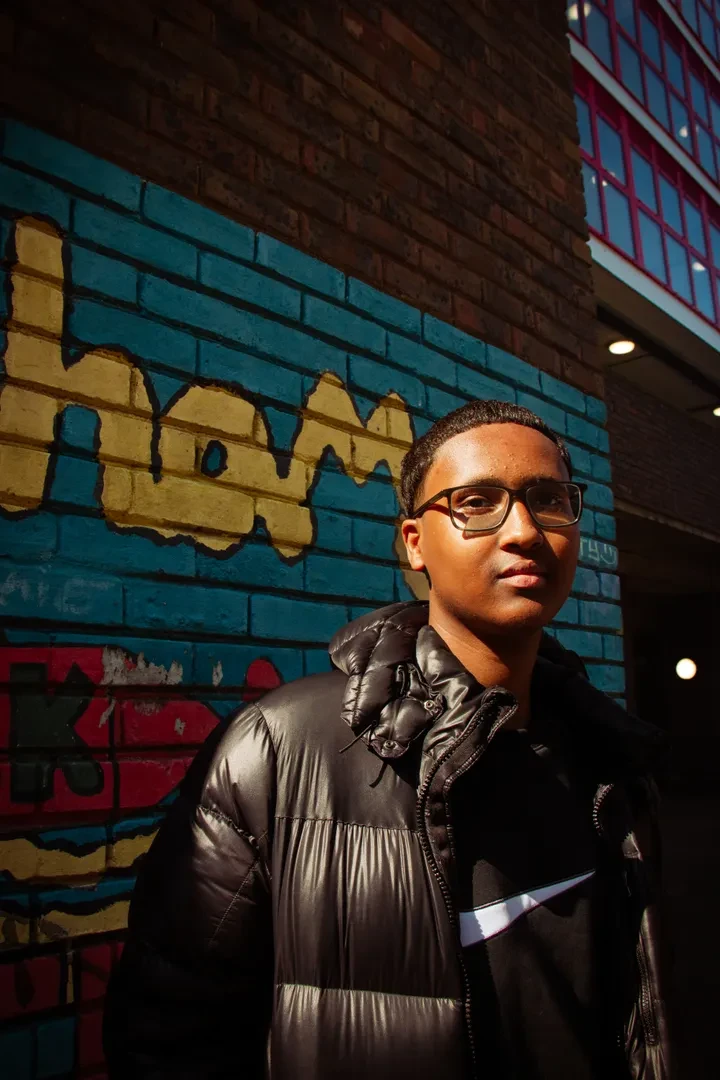 A young person of color with glasses and a neutral expressions stands in front of a graffitied brick wall, looking at the camera.