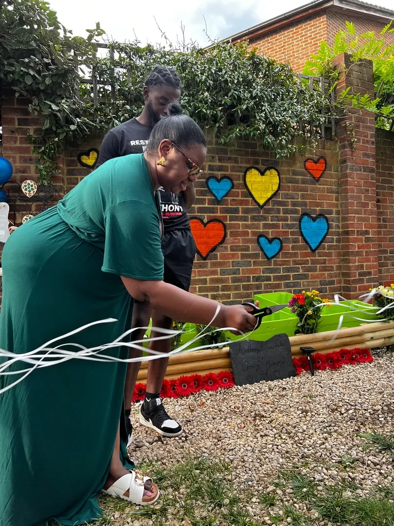 At an inauguration, a person in a formal dress cuts a ribbon with scissors, while a young person watches standing close to them. In the background, there are flowers and a memorial slate next to a brick wall with colorful heart murals.