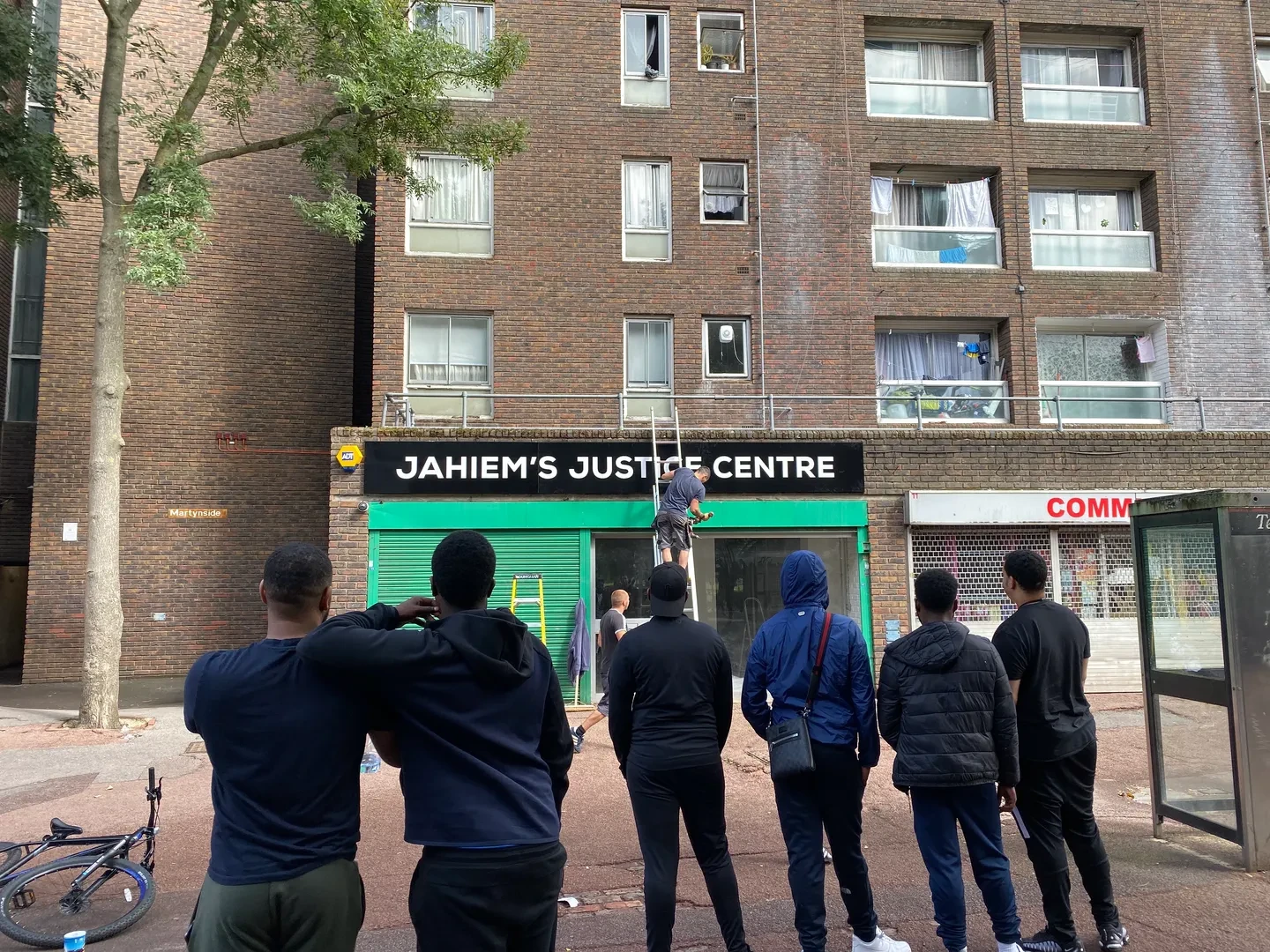 A small group of six people are standing in front of Jahiem's Justice Centre, turning their back to the camera and looking at two workers who are repairing the Centre's sign.