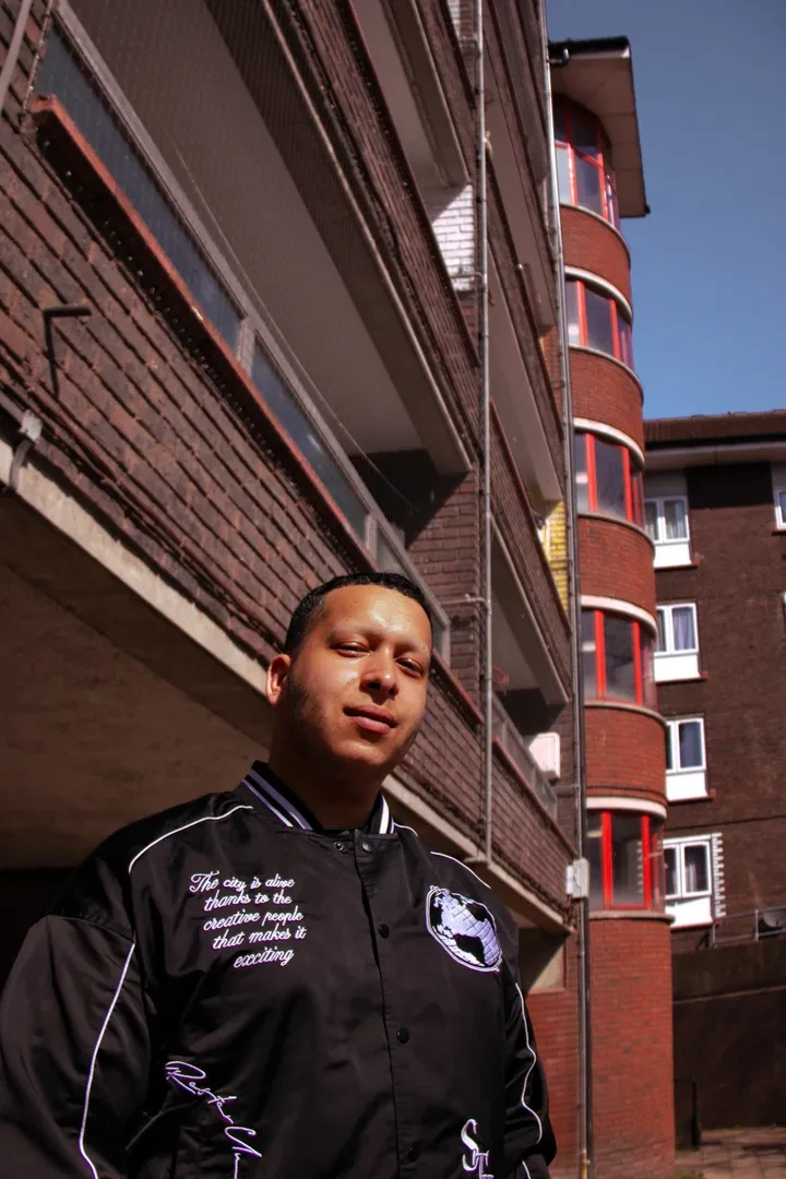 A young person is standing in front of a brick building, looking at the camera with a neutral expression.