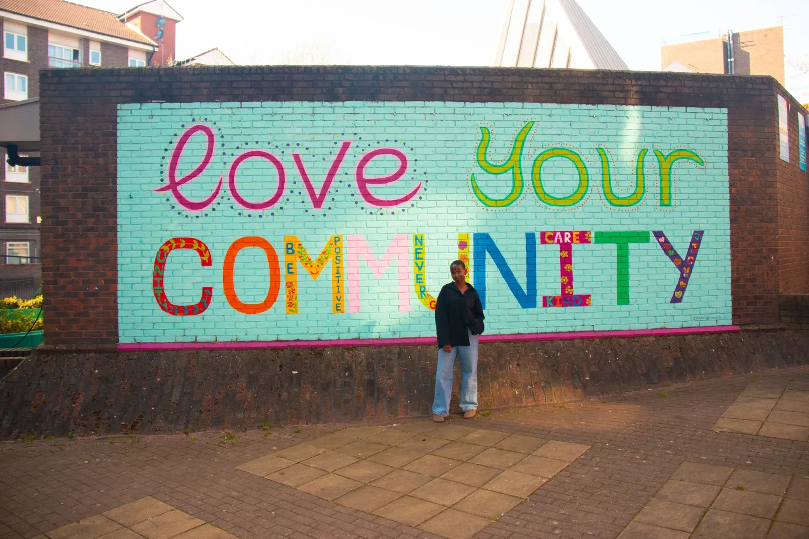 A smiling young person is standing in front of a large, colorful mural on a brick wall that reads "Love your community"