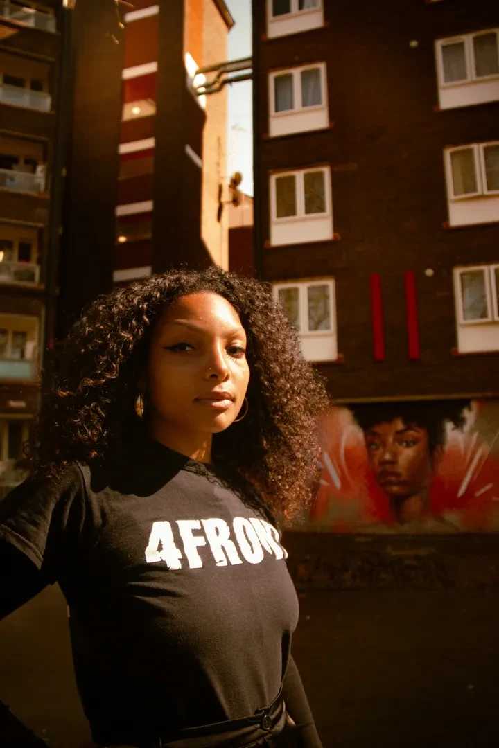 A young person with medium-length coily hair stands in front of a brick building wearing a black t-shirt with "4FRONT" written in white on it and looking at the camera.