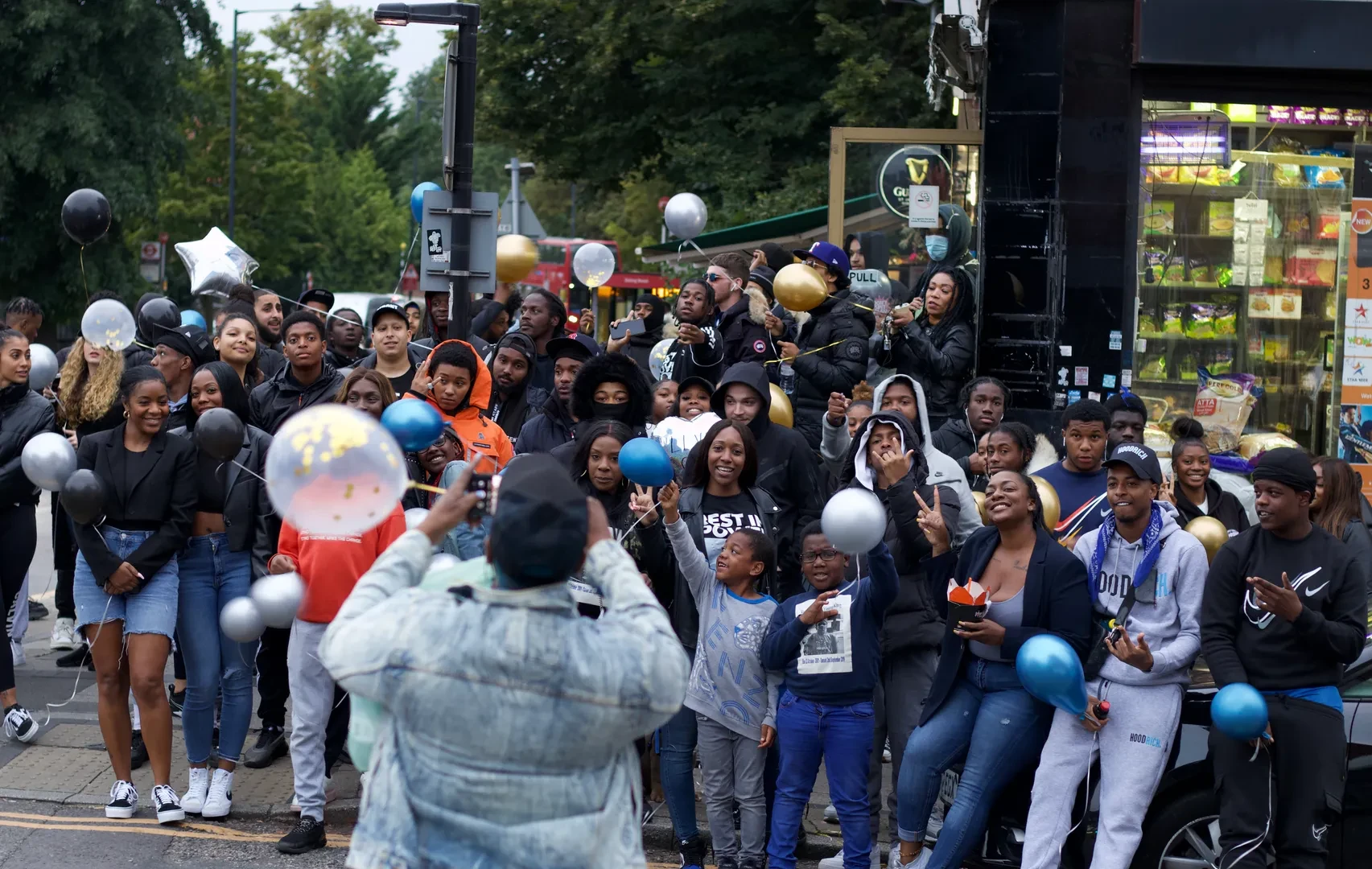 A very large group of people of color, mainly teens and young adults and a few children, are standing in an urban environment. Some of them are holding party balloons. One person is turning their back on the camera to take a picture of the gathering.